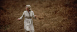a woman in a long tan coat runs in tall golden grass while looking over her shoulder. the image is grainy and warped. 